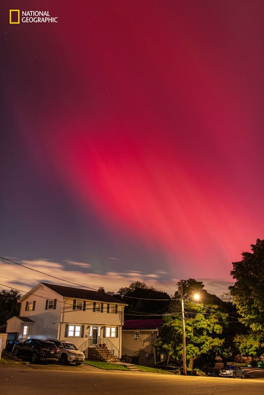 Una calle suburbana de noche, con una casa en primer plano y espectaculares auroras rojas y rosas iluminando el cielo. El indicador National Geographic aparece en la esquina superior izquierda.
