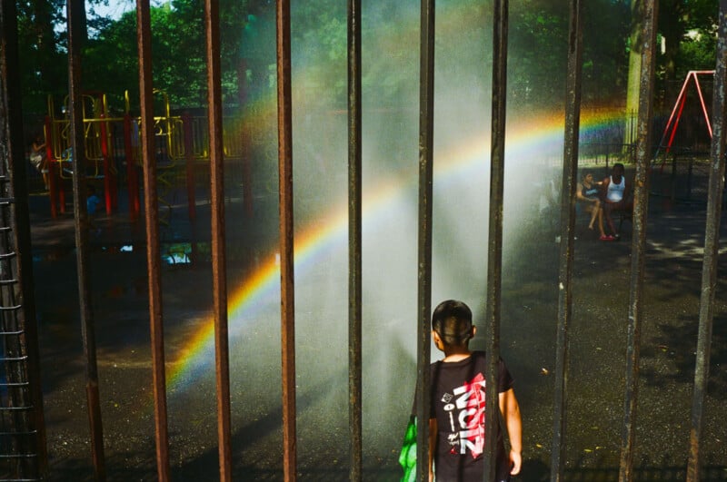 A young boy stands behind metal bars at a playground, looking at a bright rainbow formed by water mist. Two children sit on a bench in the background near playground equipment. Sunlight filters through the trees.