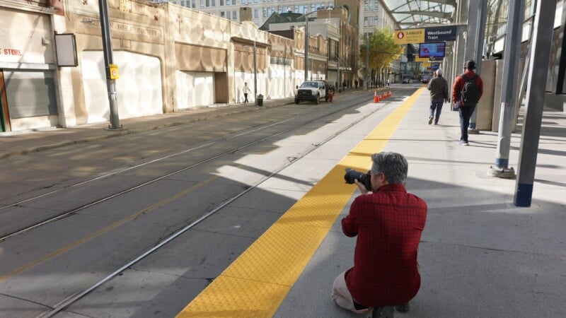 Un hombre con una camisa a cuadros rojo se arrodilla en la acera, fotografiando una tranquila calle de la ciudad y pistas de tranvía. Algunas personas vinieron de lejos, una "Tren hacia el oeste" El letrero es visible sobre la cabeza.