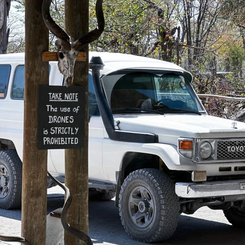 A white Toyota Land Cruiser is parked near a wooden post with a sign that reads, "TAKE NOTE: The use of DRONES is STRICTLY PROHIBITED." There’s a skull mounted above the sign and trees in the background.