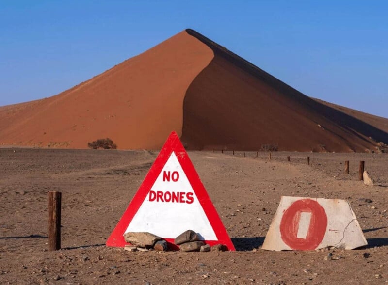A large sand dune rises in the background under a clear blue sky. In the foreground, a triangular sign with red borders reads "NO DRONES," and a stone marker with a red "O" is placed beside it.