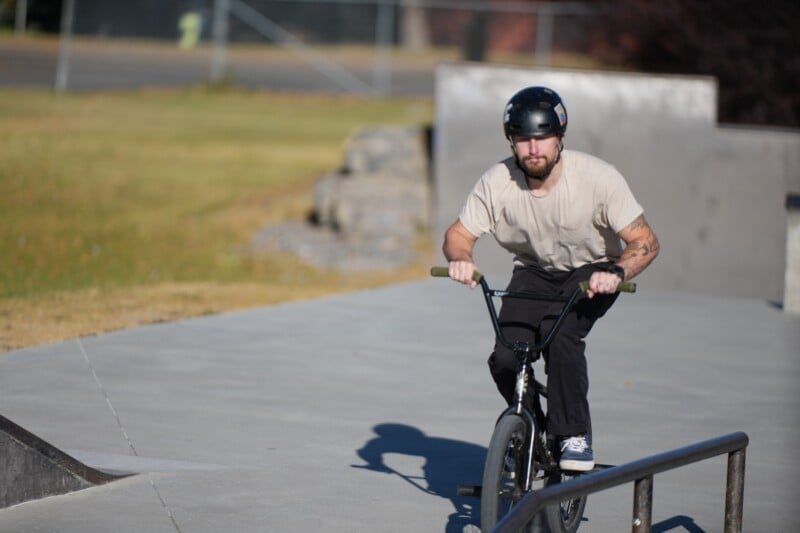 Un hombre que lleva un casco y una camisa de color claro monta una bicicleta BMX en un parque de patinaje en un día soleado, acercándose a una barandilla de metal.