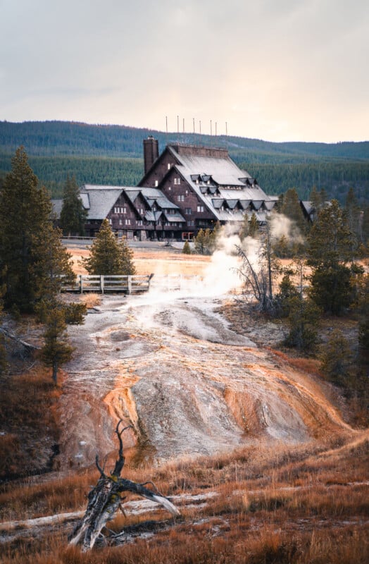 A large rustic lodge with a steep roof stands among pine trees, with steam rising from geothermal vents in the foreground and rolling hills in the background under a cloudy sky.