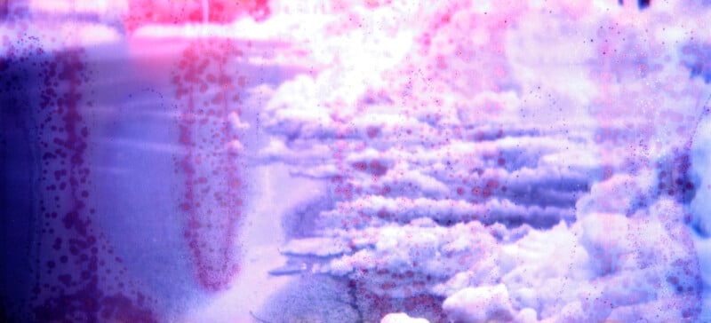 A dreamy, abstract image showing snow on the ground with pink and purple light leaks and speckled textures, creating a soft, atmospheric effect.