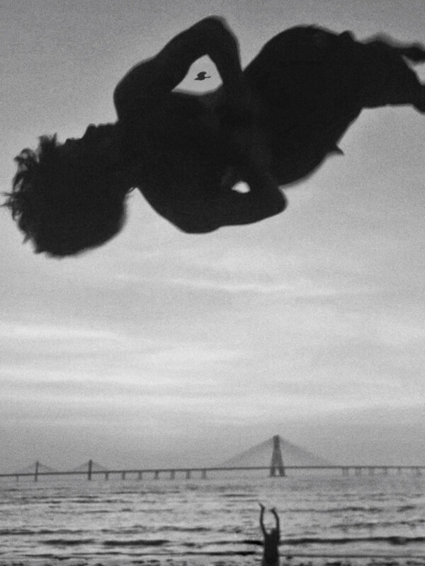 A person flips high in the air in silhouette above a beach, with another person below and a long bridge in the background at sunset. A bird flies nearby in the sky. The photo is in black and white.
