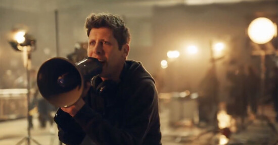 A person holding a megaphone is giving directions on a film set, surrounded by bright lights and filming equipment in a dimly lit environment.