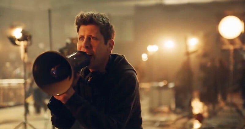 A person holding a megaphone is giving directions on a film set, surrounded by bright lights and filming equipment in a dimly lit environment.