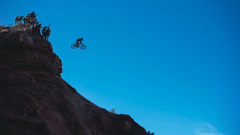 A cyclist performs a dramatic jump off a high rocky cliff, silhouetted against a clear blue sky, while a group of people on the cliff edge watch and cheer.