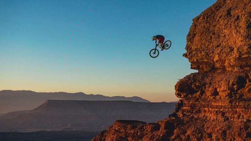 A mountain biker wearing a helmet rides off a steep rocky cliff at sunset, with dramatic desert mountains and a clear sky in the background.