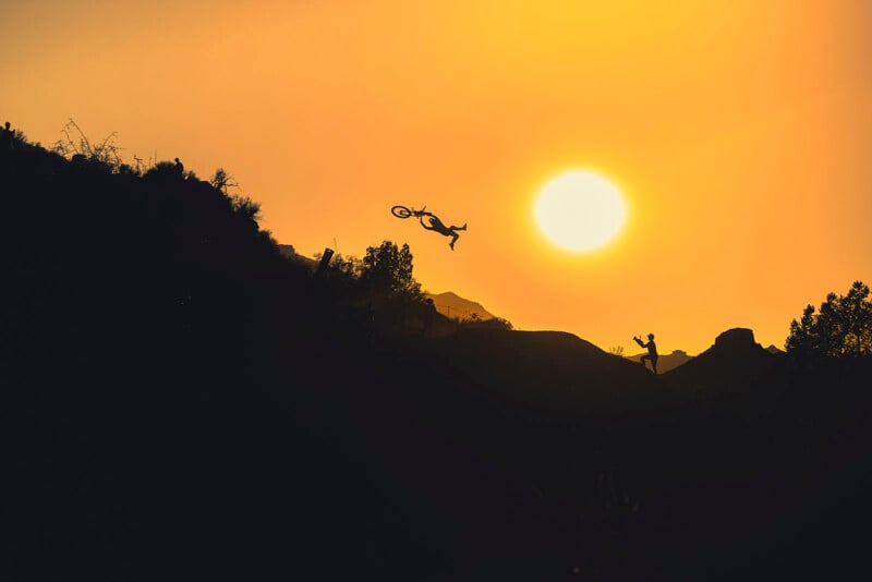 A cyclist performs a mid-air stunt on a bike against a bright orange sunset, with silhouetted hills and trees in the foreground and a person watching from below.