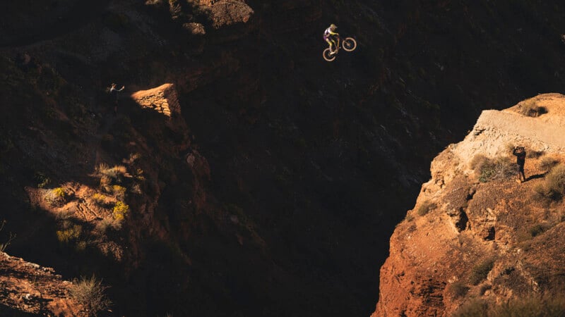 A mountain biker soars through the air over a deep canyon, with rugged red cliffs and sparse vegetation below, while a person stands on a ledge watching the jump.