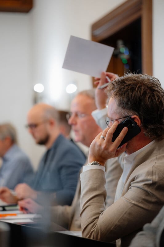A man in a beige jacket talks on the phone and raises a piece of paper during a meeting. Other people sit next to him, focused on their work. The setting appears professional and formal.