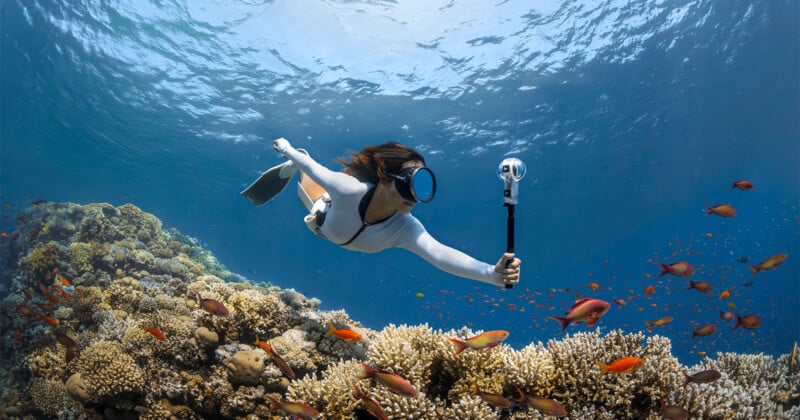 Una mujer que lleva un traje de baño blanco y una máscara de snorkel nada en un vibrante arrecife de coral sosteniendo una cámara submarina. Peces de colores la rodean mientras el sol brilla a través del agua azul clara.