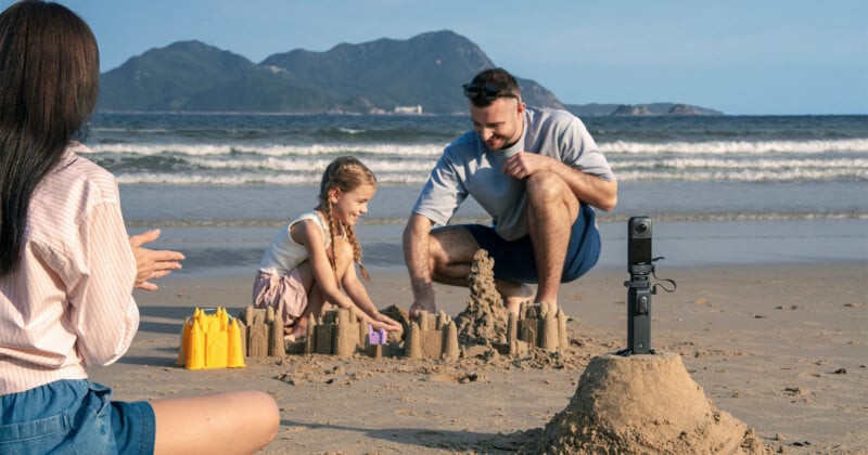 Un hombre y una niña construyen castillos de arena en la playa mientras una mujer observa y aplaude. Cerca había una cámara montada en un pequeño trípode. Al fondo se ven olas y colinas verdes.