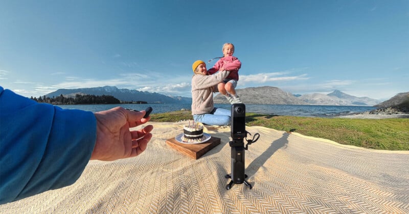 Un hombre sostiene a un niño sonriente sobre una manta de picnic cerca de un lago con montañas al fondo. Sobre la manta hay una tarta y en el otro lado un mando a distancia, cerca de la cámara montada sobre un trípode. La escena era luminosa y alegre.