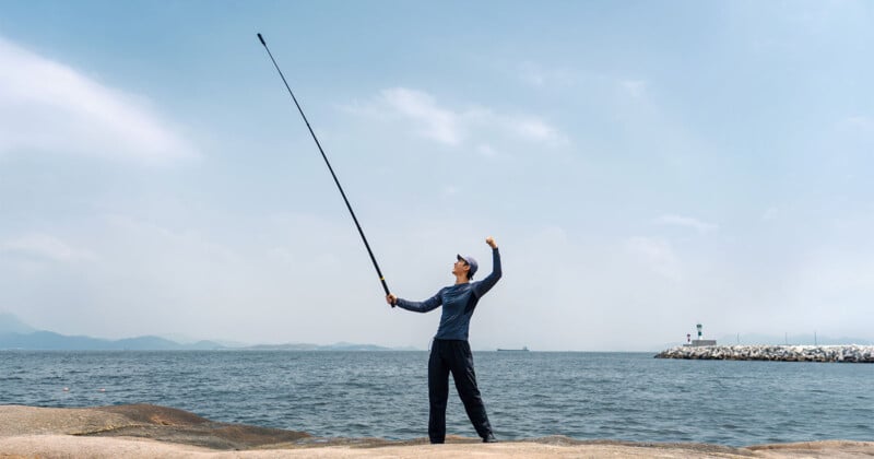 Un hombre estaba de pie sobre una roca junto al mar, sosteniendo con entusiasmo una larga caña de pescar en una mano. El cielo estaba despejado, con algunas nubes y a lo lejos se veía un faro.