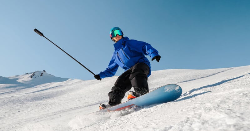 Un esquiador con una chaqueta azul y gafas reflectantes se desliza por las pistas en un día soleado sosteniendo un palo selfie con una cámara. El fondo son montañas nevadas y un cielo azul claro.
