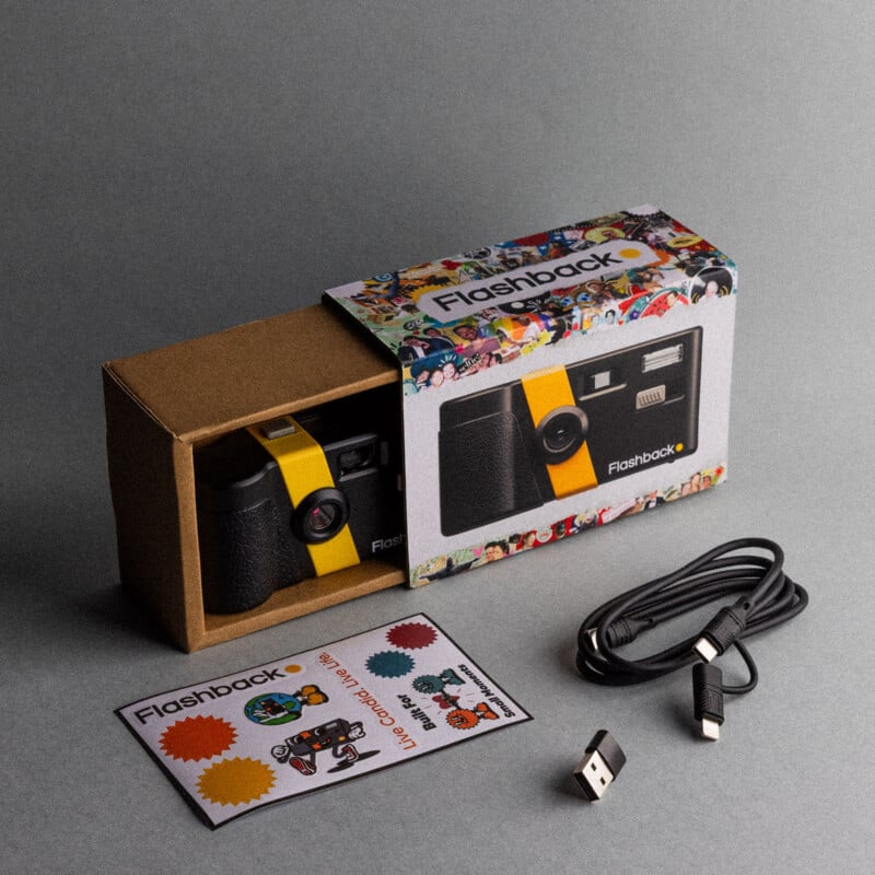 A boxed camera labeled "Flashback" partially slid open, revealing the camera inside. Nearby are a USB cable, a USB adapter, and a colorful instruction card on a gray surface.