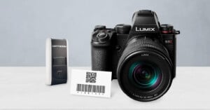 A Lumix S5 II camera, an Opticon barcode scanner, and a card displaying a QR code with a barcode, all placed on a white surface against a light gray background.