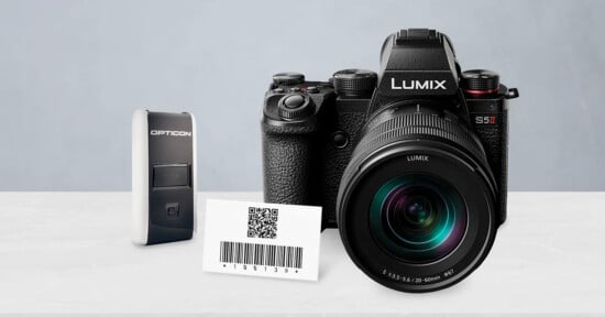 A Lumix S5 II camera, an Opticon barcode scanner, and a card displaying a QR code with a barcode, all placed on a white surface against a light gray background.