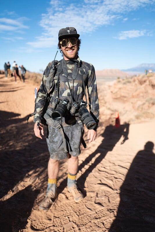 A man wearing camouflage clothing, sunglasses, and a hat stands on a dusty trail with two cameras around his neck. The background shows people walking, red rock terrain, and a blue sky with scattered clouds.