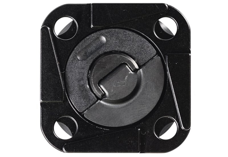 Primer plano de un pestillo o sujetador empotrado de metal negro con un centro redondo y cuatro orificios para tornillos en las esquinas, montado sobre una placa de metal cuadrada.