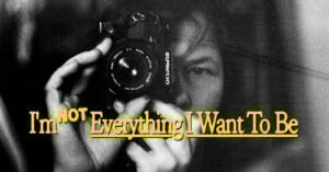 A person holds a camera close to their face, partially obscured in black and white. Yellow text over the image reads, "I'm NOT Everything I Want To Be.