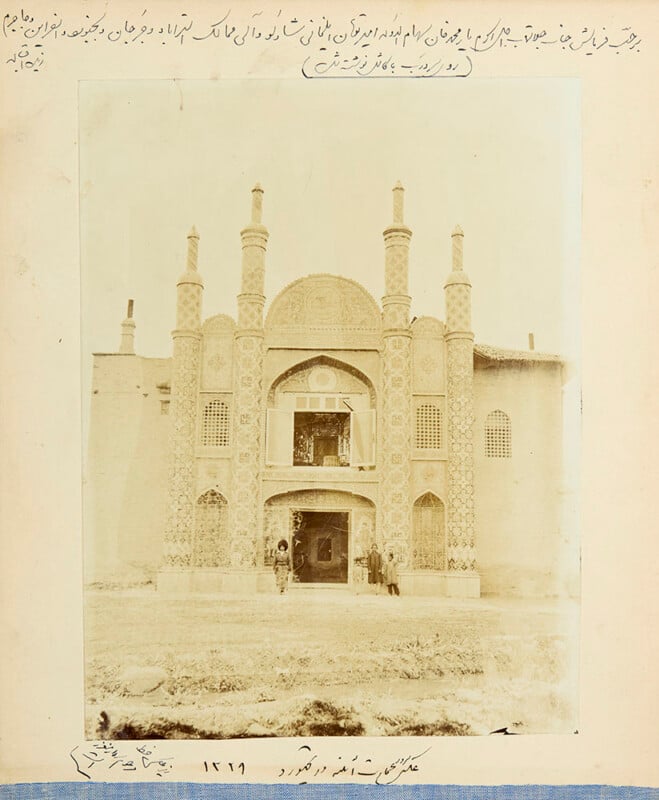 Fotografía en sepia de un edificio histórico persa con ladrillos ornamentados, arcos y cuatro torres; varias personas paradas en la puerta. Hay texto persa escrito a mano que rodea la imagen.