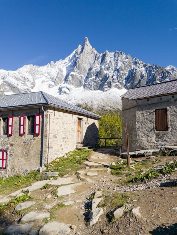Two stone mountain huts with red-trimmed windows stand on a rocky path, with a dramatic, snowy jagged peak rising sharply in the background under a clear blue sky.