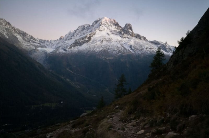 A snow-covered mountain peak rises above a dark, forested valley at dusk. Rocky slopes and pine trees line the foreground, with a winding path leading toward the majestic mountain under a clear sky.