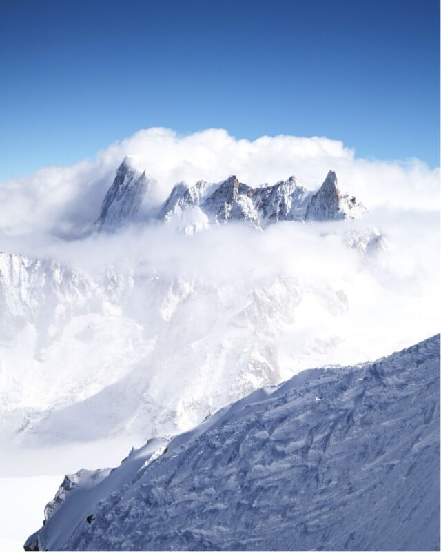 Snow-covered mountain peaks rise above clouds under a clear blue sky. The sunlight highlights the rugged, rocky summits, while mist and snow blanket the lower slopes, creating a dramatic and serene winter landscape.