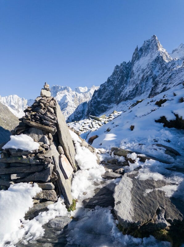 A stone cairn stands on a snowy mountain trail, surrounded by rocks and patches of ice, with rugged, snow-covered peaks rising sharply under a clear blue sky.