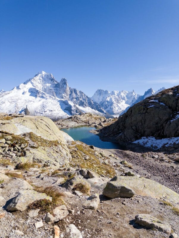 Snow-capped mountains rise behind a clear blue alpine lake surrounded by rocky terrain and patches of sparse vegetation under a bright, cloudless sky.
