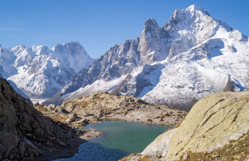 A clear blue alpine lake surrounded by rocky terrain and high snow-covered mountains under a bright blue sky.