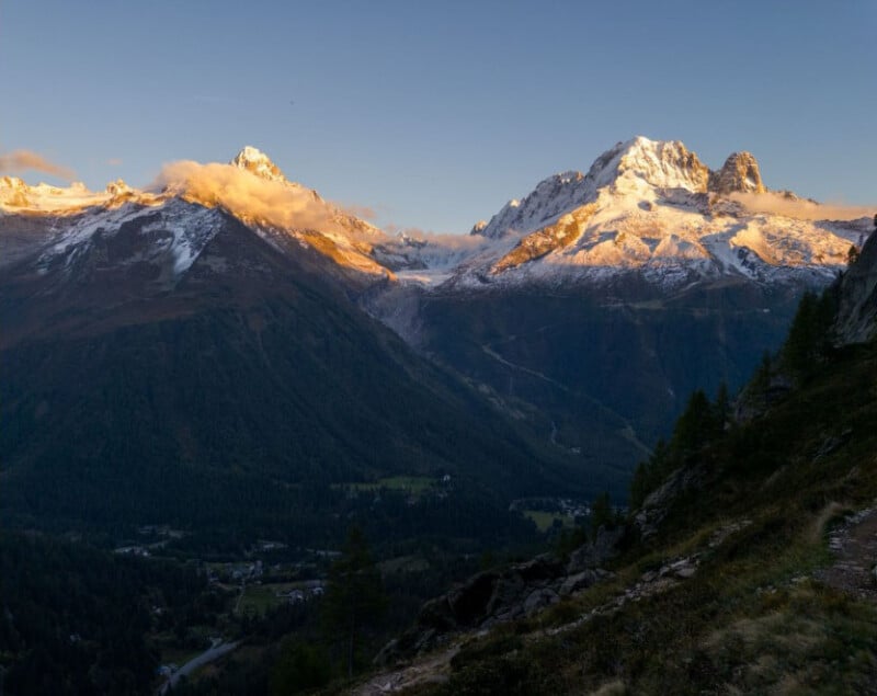 Snow-capped mountains are illuminated by the golden light of sunrise or sunset, with a green valley and small village below. Wispy clouds partially cover the peaks under a clear sky.