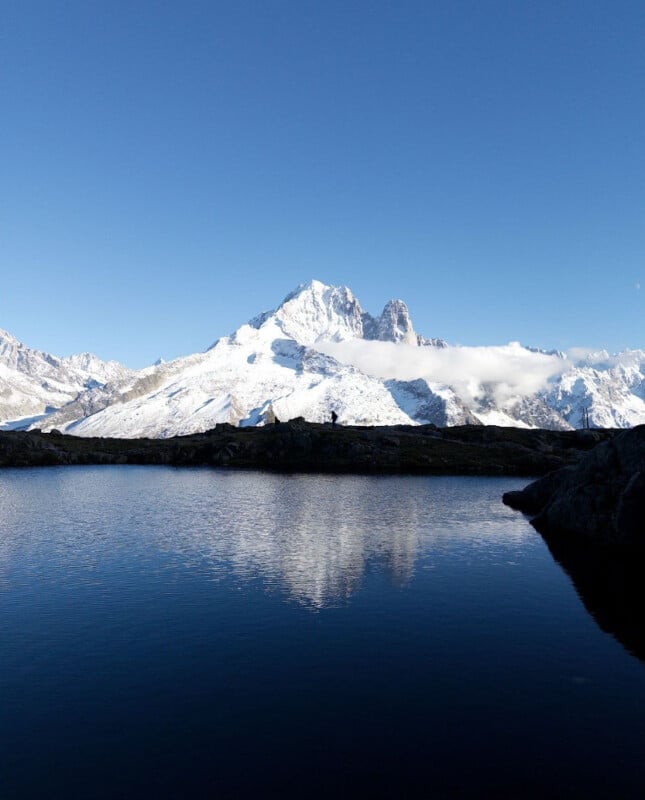 A snow-covered mountain is reflected in a calm lake under a clear blue sky, with a person standing on the shore in the distance. Wisps of clouds partially cover the mountain's base.