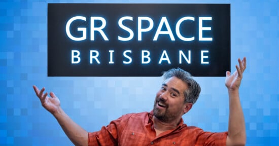 A smiling man in an orange shirt stands in front of a blue pixelated background, gesturing with raised hands beneath a sign that reads "GR SPACE BRISBANE" in large white letters.