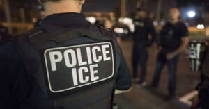 A person wearing a vest labeled "Police ICE" stands in the foreground at night, with several other people blurred in the background under streetlights.