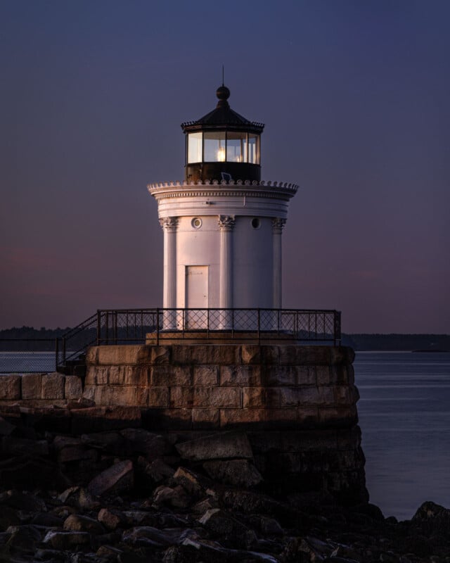 A white lighthouse with a lit lantern stands on a stone pier at dusk, surrounded by calm water and rocks, with a soft twilight sky in the background.