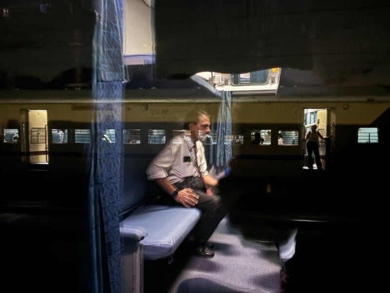 A man in formal attire sits on a blue train bunk, seen through a window with reflections. The train interior is dimly lit, with blue curtains and yellow walls, and another person is visible in the background near an open door.