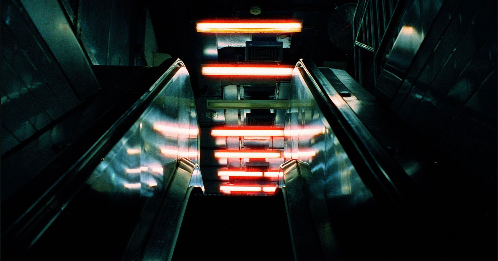 View from the top of a dark escalator descending, with bright red and white horizontal lights reflecting off the metal surfaces, creating a futuristic and moody atmosphere.