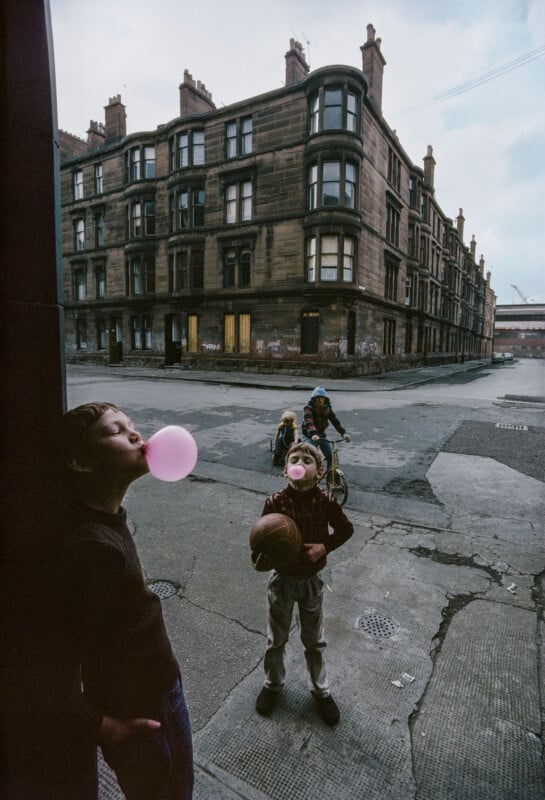Two children in the foreground blow pink bubblegum bubbles, one holding a basketball. In the background, two more kids ride bicycles near an old apartment building on an empty city street.