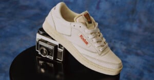 A cream-colored sneaker with "Kodak" branding rests on top of a vintage Kodak camera, set against a blue textured background.
