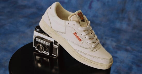 A cream-colored sneaker with "Kodak" branding rests on top of a vintage Kodak camera, set against a blue textured background.