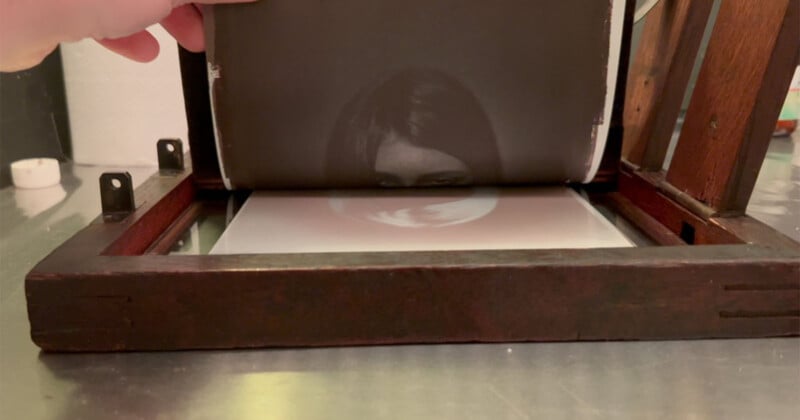 A hand lifts a sheet with a black-and-white image of a woman’s face from a wooden photo printing frame on a reflective metal surface.