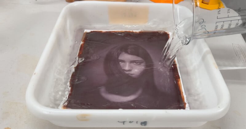 A black-and-white photograph of a woman’s face is soaking in a white tray of liquid, while water is being poured onto it from a beaker, likely as part of a photo development process.