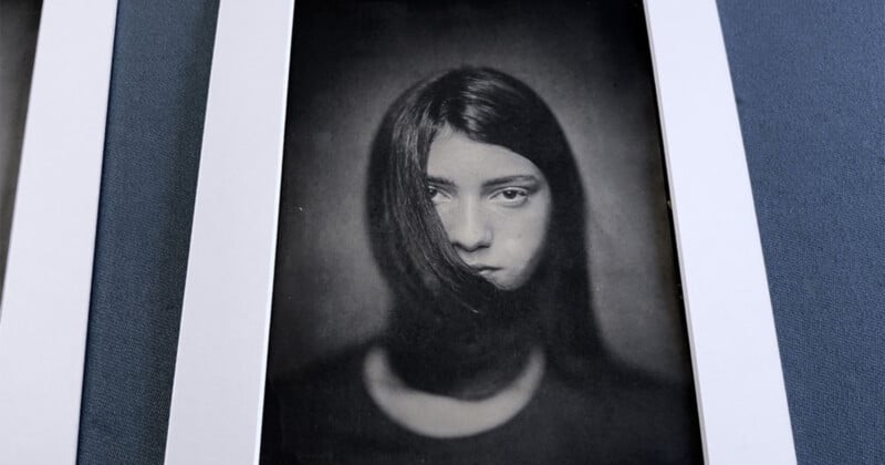 A black and white portrait of a woman with long hair partially covering her face, framed by a white border and set against a textured, blue-gray surface.