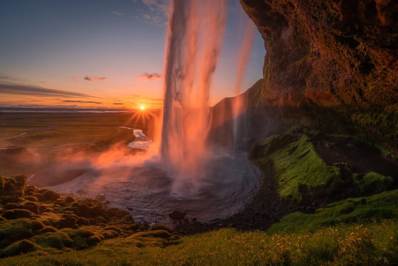 Una vibrante puesta de sol ilumina una alta cascada que cae sobre un acantilado cubierto de musgo hacia una piscina debajo, donde la exuberante hierba verde y los campos se extienden en la distancia bajo un cielo colorido.