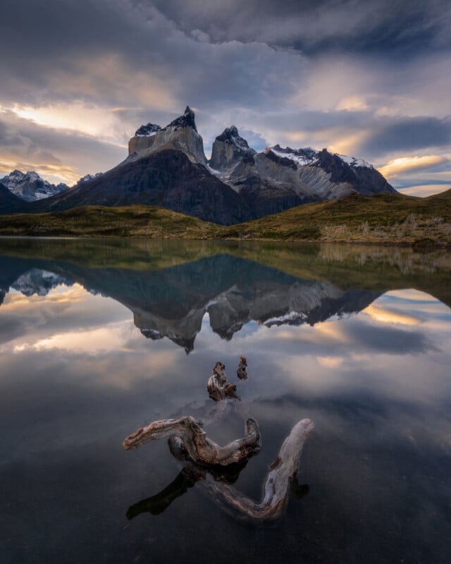 Al atardecer, las magníficas montañas se reflejan en el tranquilo lago, con madera flotante en el agua en primer plano y nubes llenando el cielo.