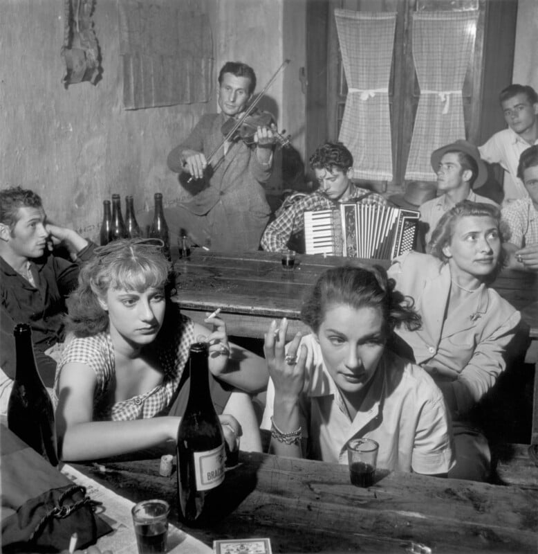 A black and white photo of a group of people sitting at wooden tables in a dimly lit bar; some hold drinks, while two musicians in the background play violin and accordion. The mood is lively and social.